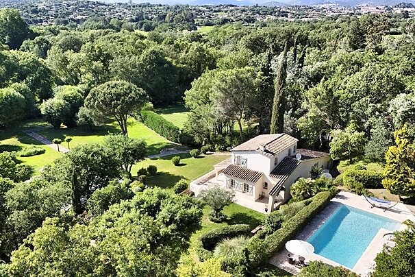 Aerial view of a charming villa with a sparkling pool, nestled in a sprawling green landscape surrounded by lush trees and distant towns.
