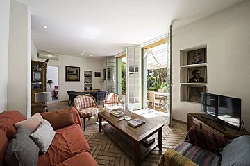 A warm living room with a herringbone tile floor, red sofa, and French doors opening to a garden. A TV sits by recessed portraits on the wall.