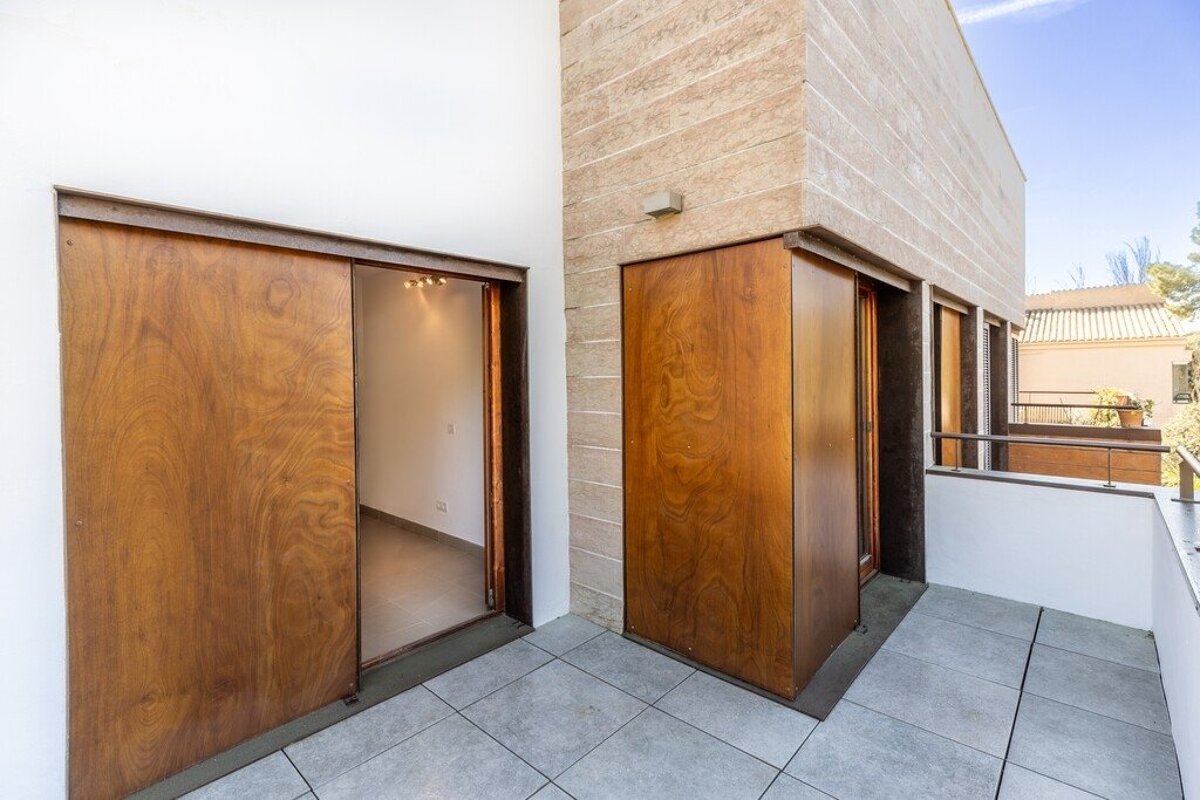 Modern balcony with grey tiled floor, sliding wooden doors, and white and beige stone walls under a blue sky.