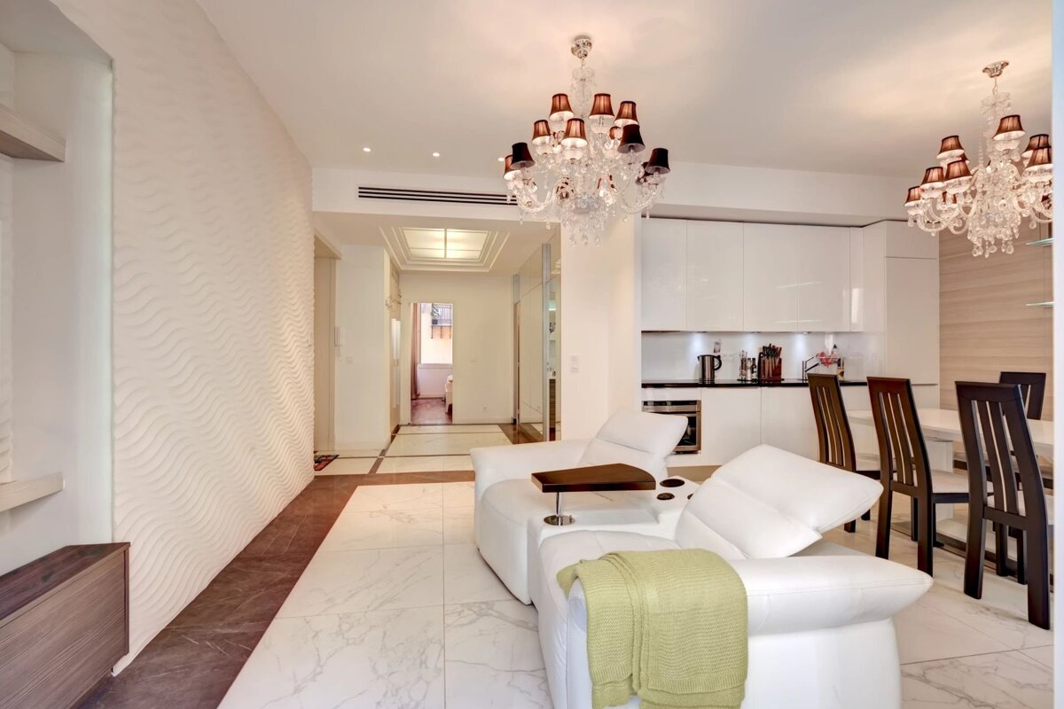 A luxurious modern open-plan living room, kitchen & dining. Features white leather sofas, elegant chandeliers, a textured wall, and marble-look tiles.