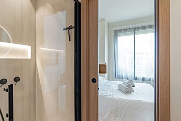View from a sleek bathroom with glass shower into a bright bedroom with a white bed, rolled towels, and a window, framed by a wooden doorway.