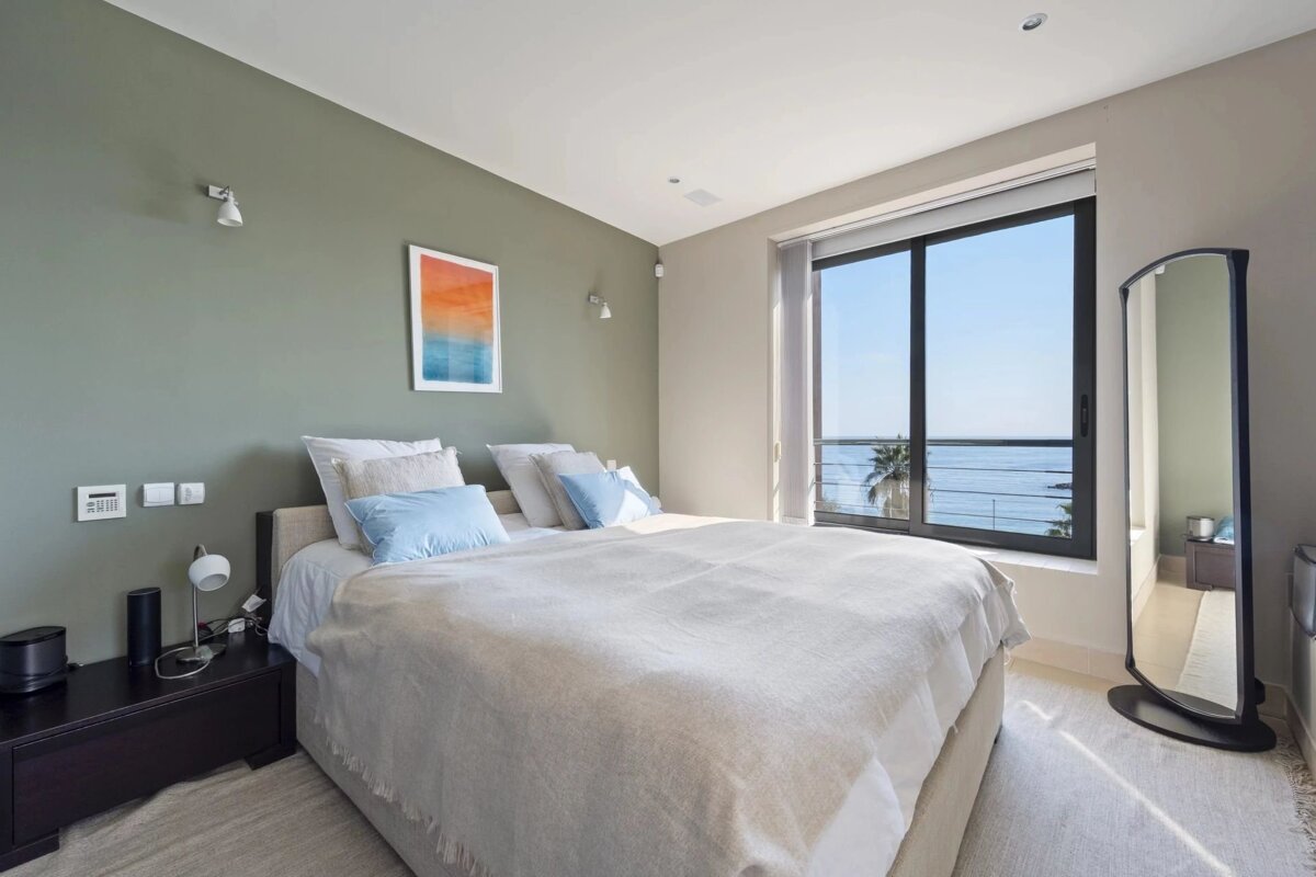 A modern bedroom with a sage green accent wall, light bedding, and a large window overlooking the ocean and a palm tree.