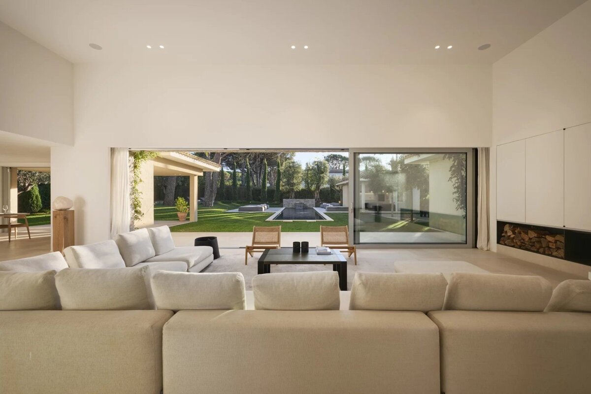 A bright, minimalist living room featuring a large sectional sofa and glass doors opening to a lush backyard with a pool.
