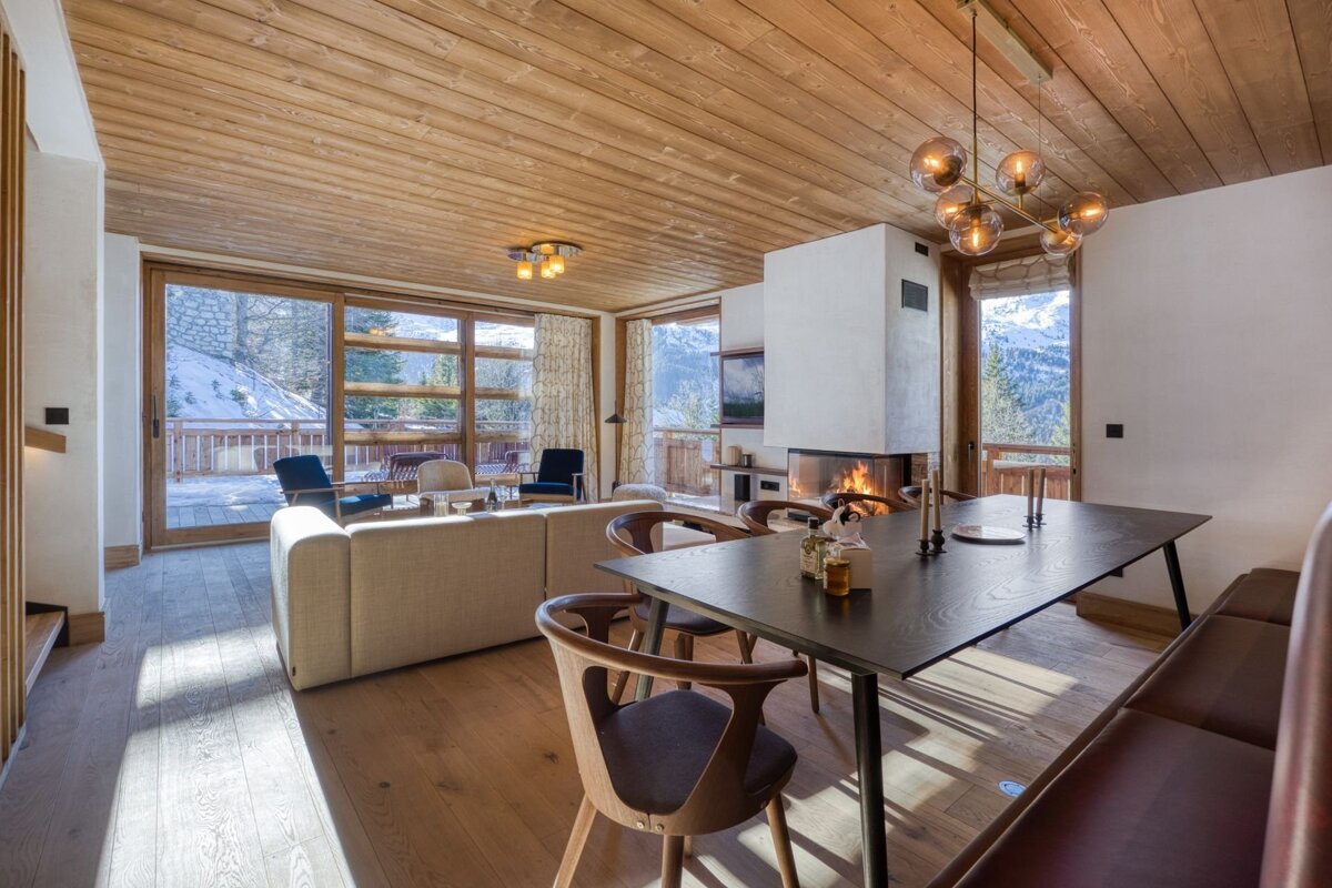 A luxurious open-plan chalet living/dining room with a glowing fireplace, warm wood finishes, and large windows revealing a stunning snowy mountain view.
