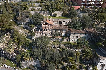 Aerial view of an old villa complex built into a lush, terraced hillside with stone walls, overgrown greenery, and palm trees.