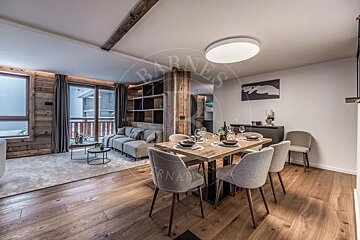 A modern open-plan living and dining area in a chalet, featuring wood paneling, a set dining table, and a cozy sofa, with snowy views outside.