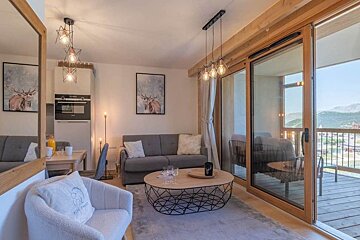 Cozy modern living room with a grey sofa, wooden accents, pendant lights, and sliding doors opening to a balcony with scenic views.