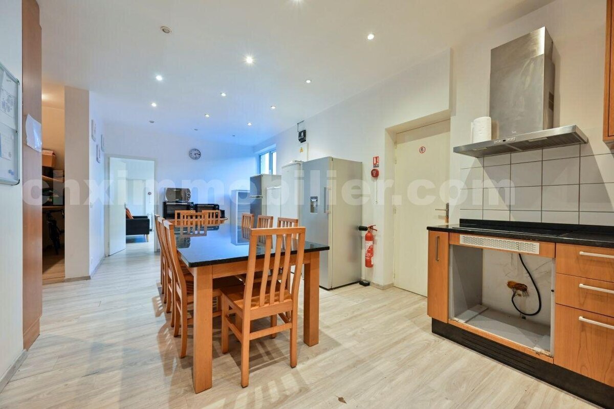 A spacious modern kitchen/dining area features a long table with chairs, multiple refrigerators, and a partially equipped kitchen counter with a range hood.