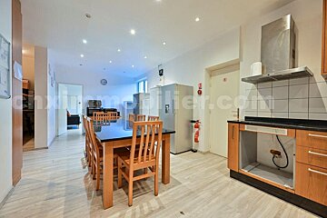 A spacious modern kitchen/dining area features a long table with chairs, multiple refrigerators, and a partially equipped kitchen counter with a range hood.