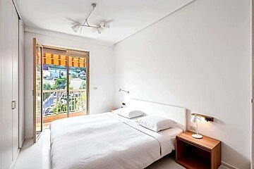 A bright, minimalist white bedroom with a bed, modern lighting, and a large balcony door overlooking a town with trees and buildings.