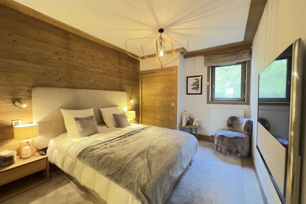 A cozy, modern bedroom features a large bed, wood-paneled accent wall, unique chandelier, sliding wood closet door, and a window.