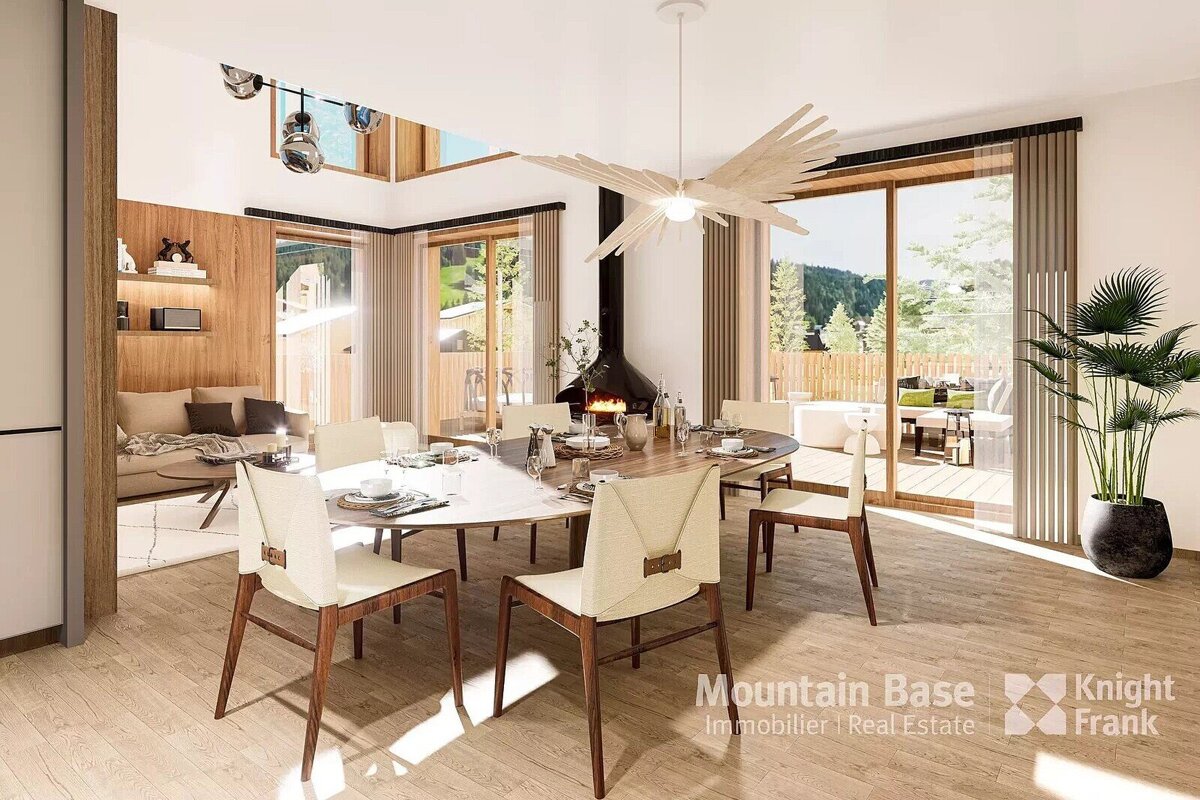 A modern, open-plan dining and living room features a large table, contemporary fireplace, and big windows looking out to nature. Bright, warm, and inviting.