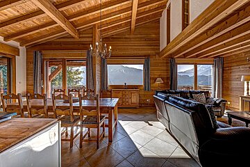 Cozy wooden chalet interior with dining and living areas, featuring large windows offering panoramic mountain views and a tiled floor.