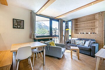 Cozy alpine apartment interior with a wooden dining table, grey sofa, armchair, and large windows showing scenic mountain views.