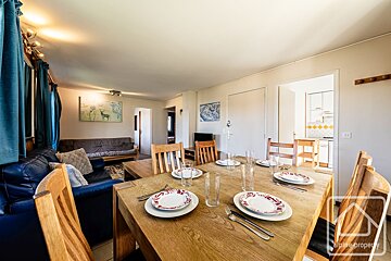A spacious open-plan living/dining room features a long wooden table set for a meal, a blue leather sofa, and a cozy futon. A kitchen is visible beyond.