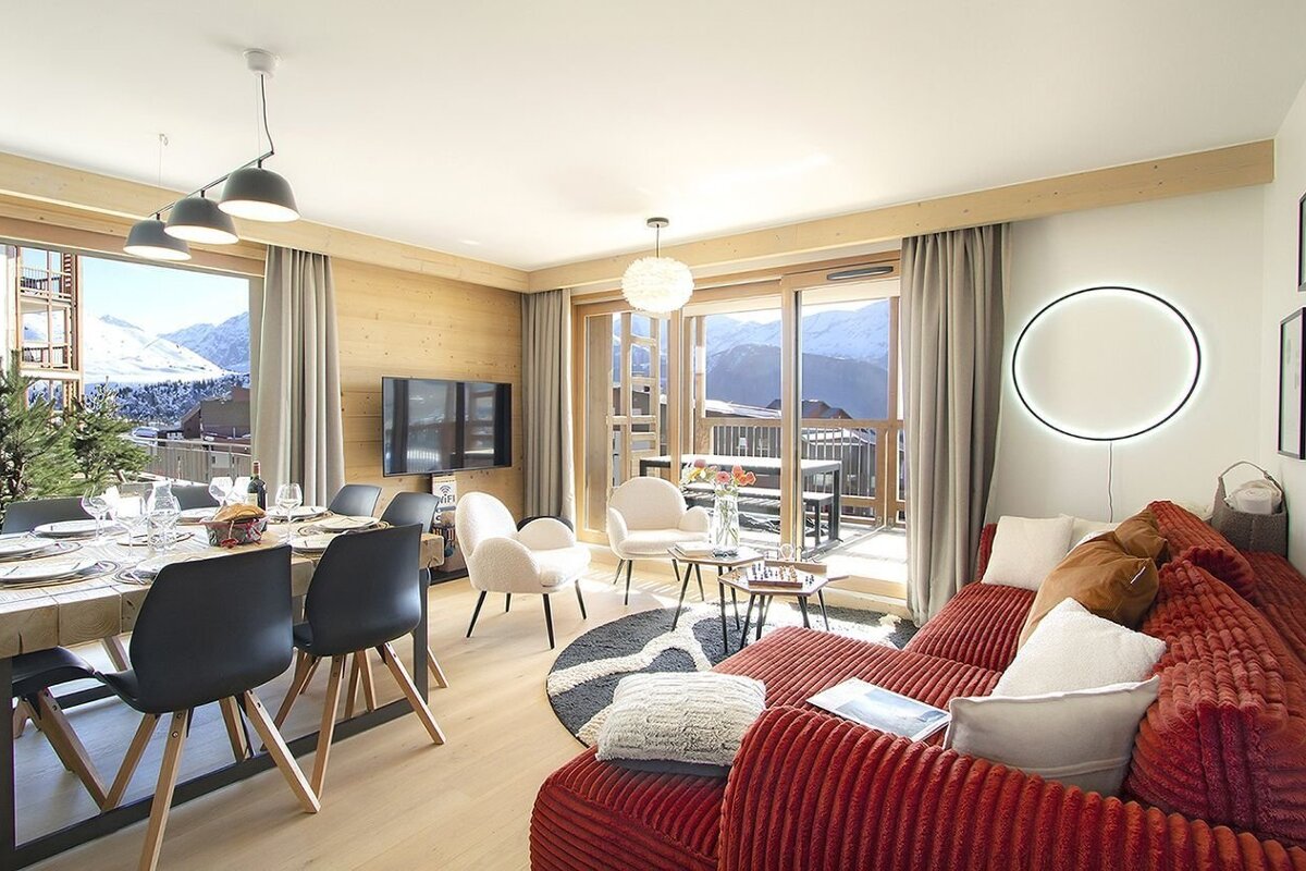 A cozy, modern chalet living/dining room with a red sofa, wood details, and large windows revealing an expansive snowy mountain view.