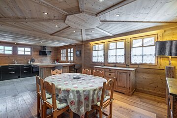 A cozy, rustic wooden chalet kitchen and dining room. Features a heart-patterned tablecloth, modern black kitchen units, and charming windows.