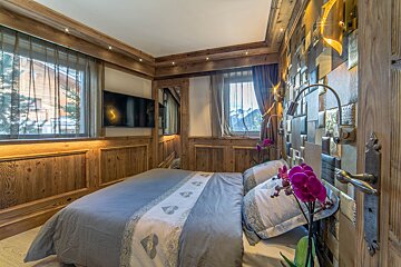 A cozy, wood-paneled bedroom with a large bed, wall-mounted TV, mountain views from windows, and a vibrant purple orchid.