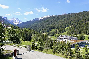 A scenic alpine valley featuring lush pine forests, resort buildings, and snow-capped mountain peaks under a clear blue sky.