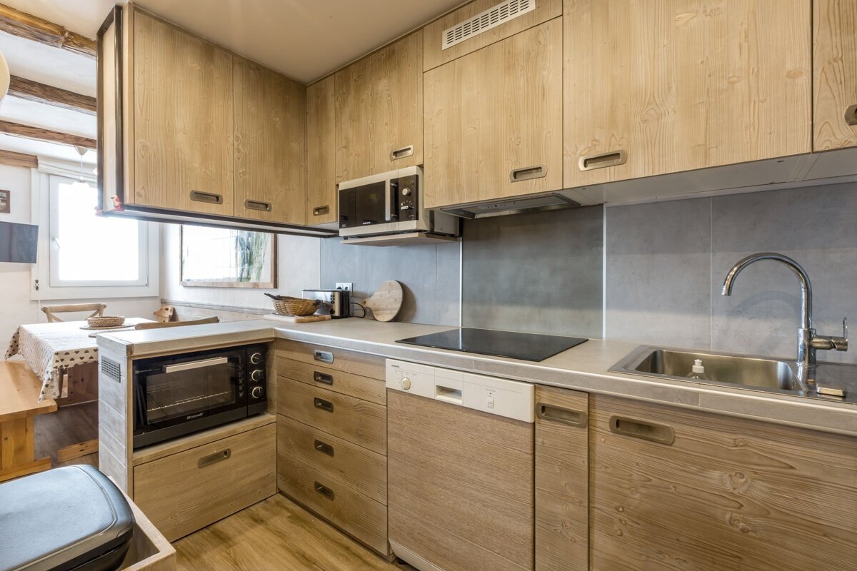 A modern kitchen with light wood cabinets, grey countertops, and integrated appliances like a microwave, oven, and sink. A dining area is visible.