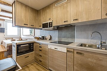 A modern kitchen with light wood cabinets, gray counters, and integrated appliances like a microwave, oven, dishwasher, stove, and sink. Dining area in background.