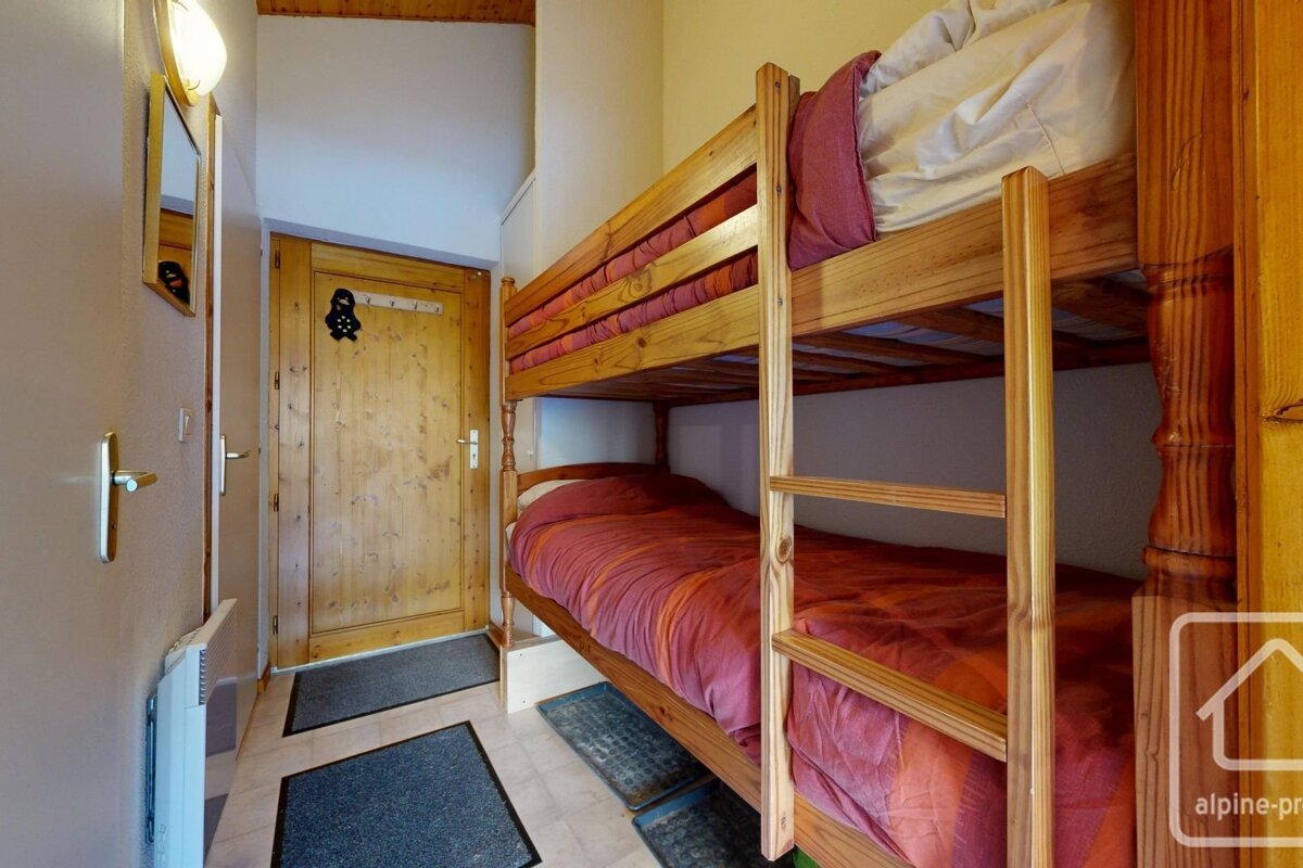 A cozy hallway with wooden bunk beds featuring red/orange bedding. A wooden door, mirror, and light fixture are also visible, creating a compact sleeping area.
