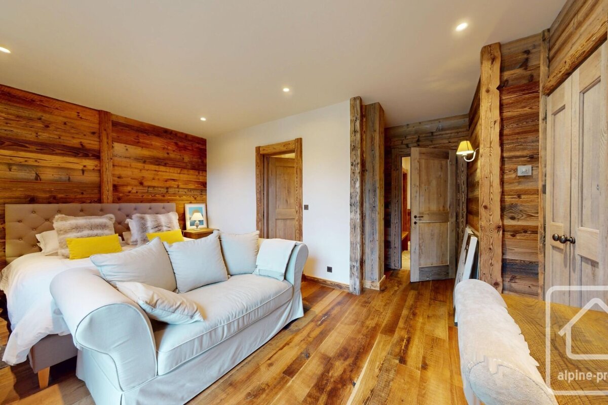 A cozy, rustic bedroom with warm wooden walls, floor, and doors. A large bed with yellow accents and a light gray sofa furnish the welcoming space.