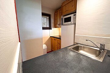 A compact kitchenette features wooden cabinetry, a microwave, fridge, and a sink set in a dark countertop. A window with blinds is visible.