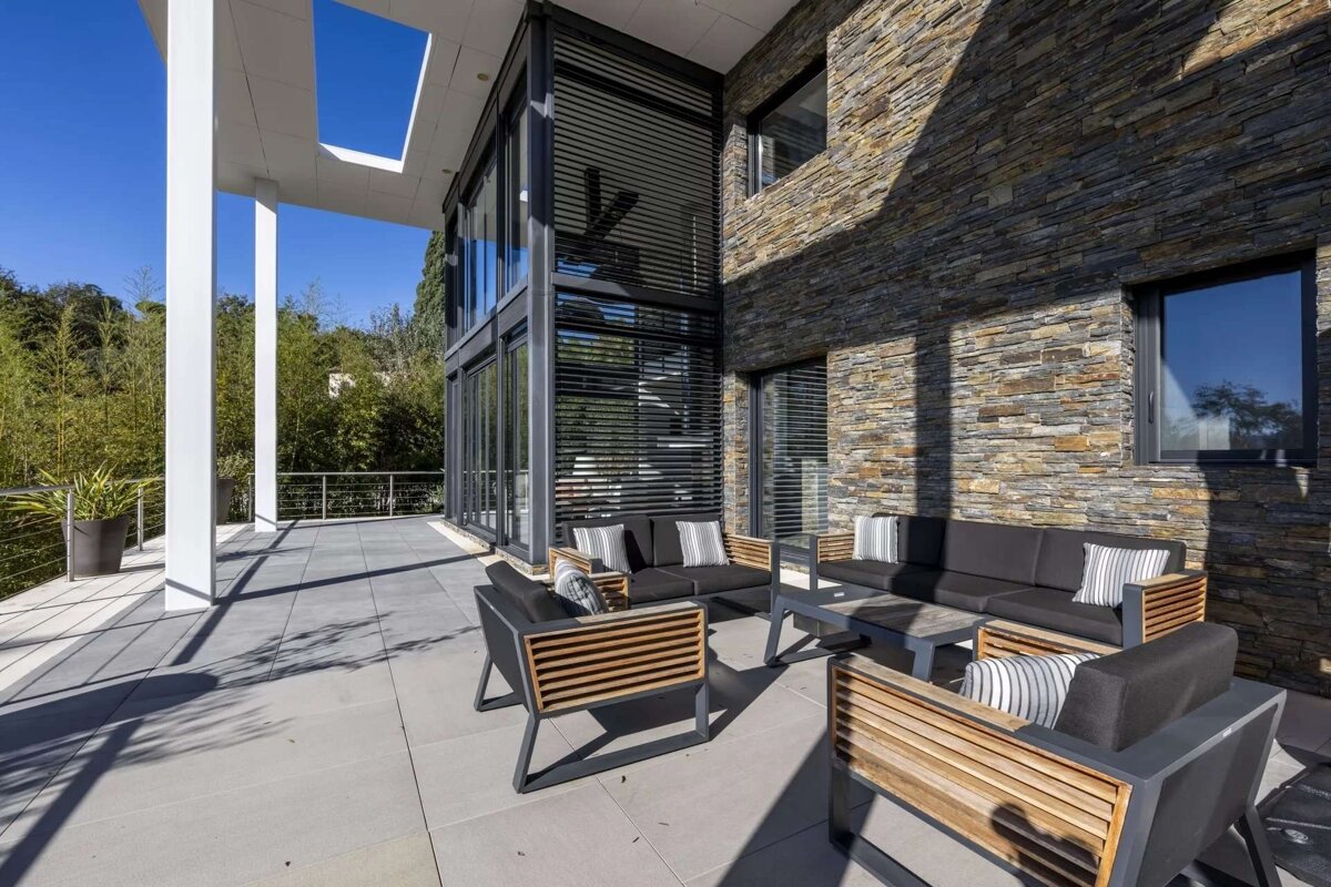 Sunny modern house patio with sleek outdoor furniture, stone walls, large windows, and lush green bamboo.