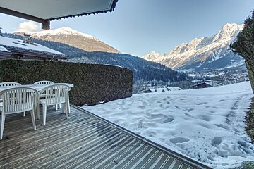 A snowy deck with a table and chairs and mountains in the background