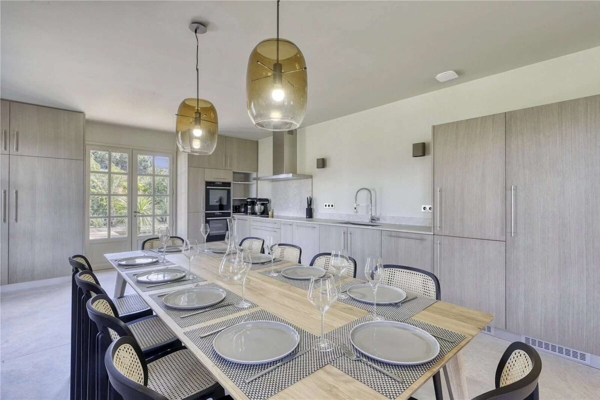 A modern kitchen and dining room with a long wooden table set for ten. Features include light gray cabinets, large windows, and two amber pendant lights.