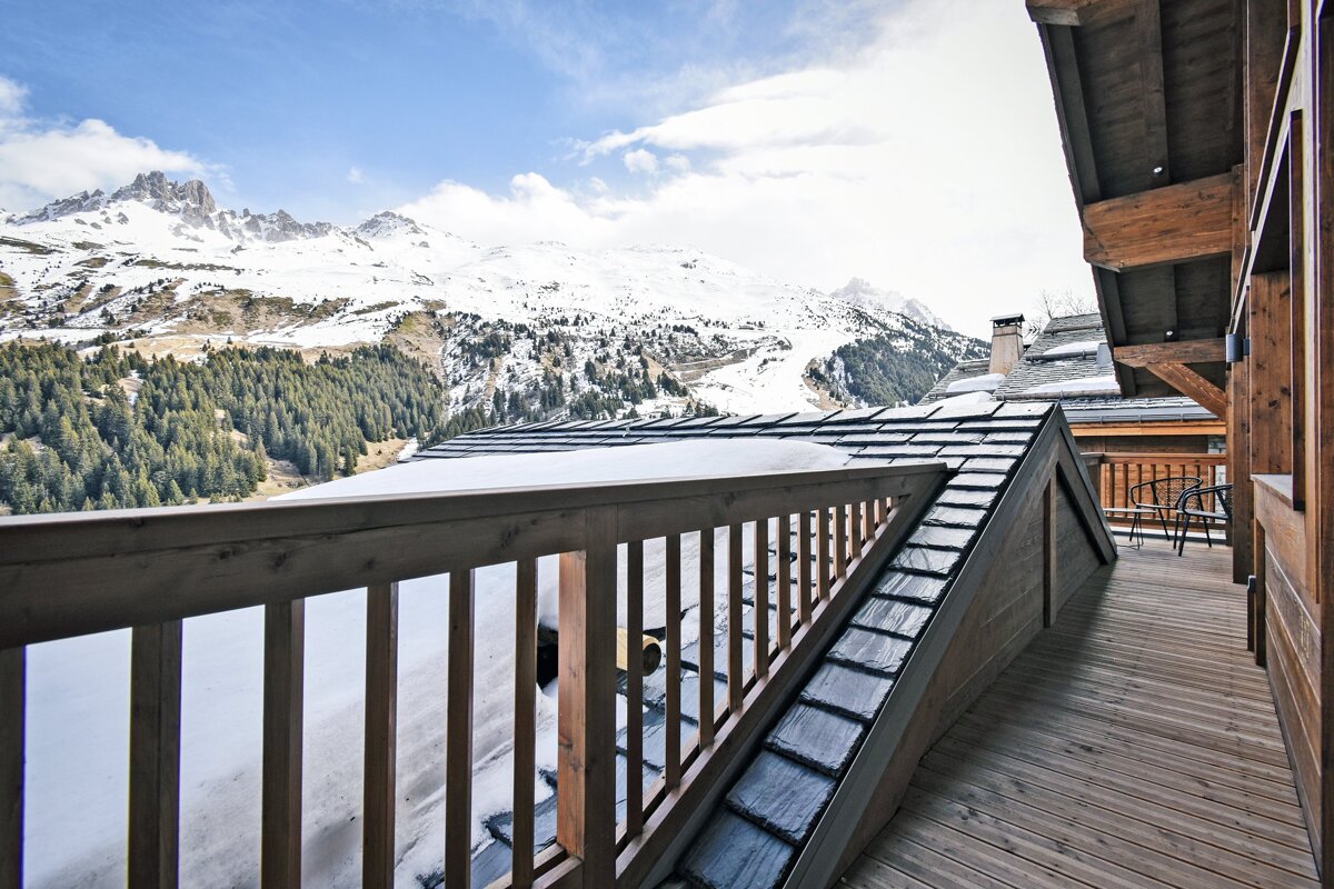 A view from a wooden chalet balcony overlooking snow-covered mountains and a pine forest under a blue sky.