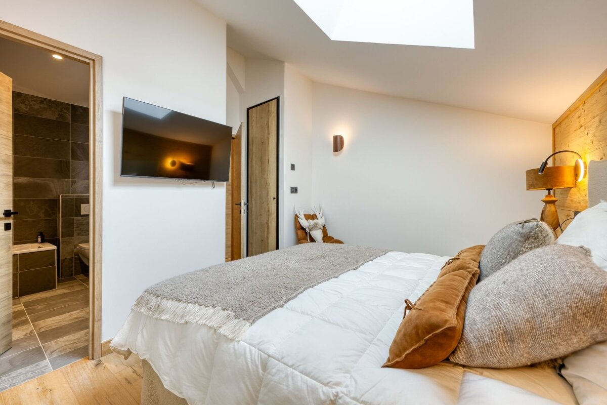 A stylish bedroom featuring a large bed with neutral bedding, a wall-mounted TV, and an open ensuite bathroom. Skylight and warm lighting create a modern, cozy feel.
