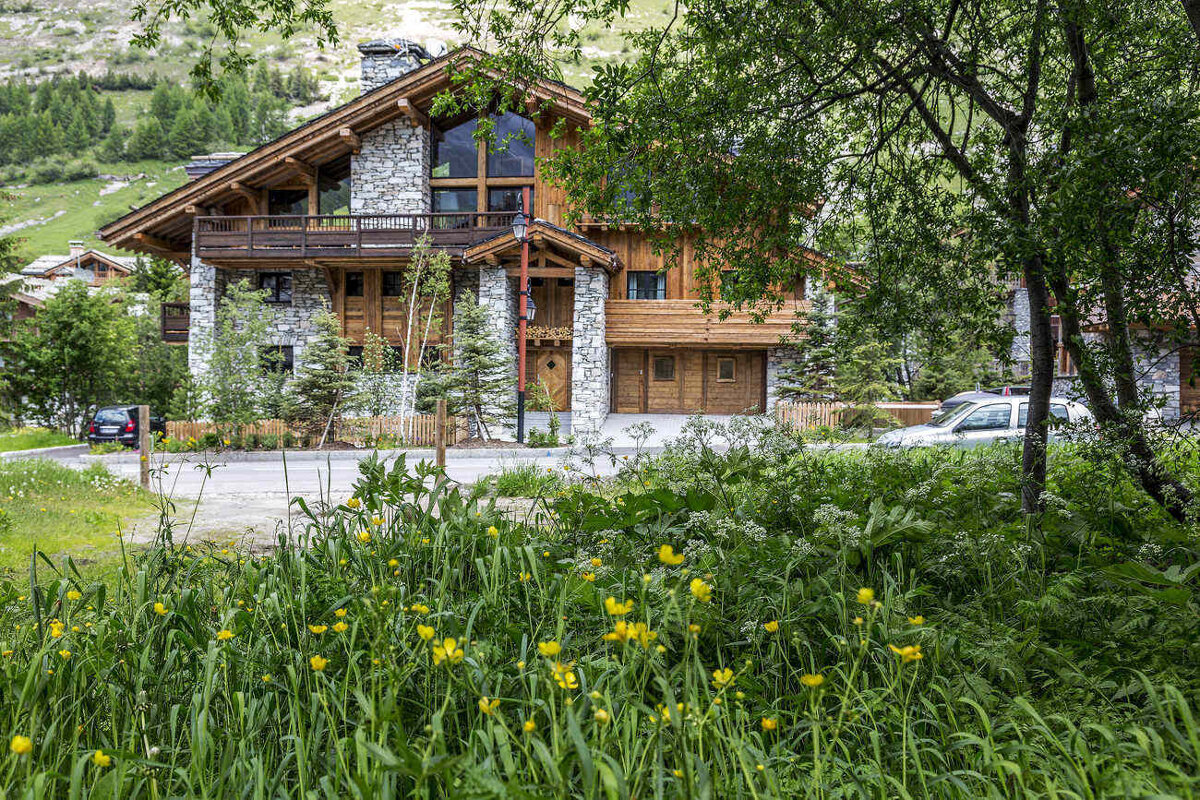 A large stone house with a wooden roof is surrounded by tall grass and trees