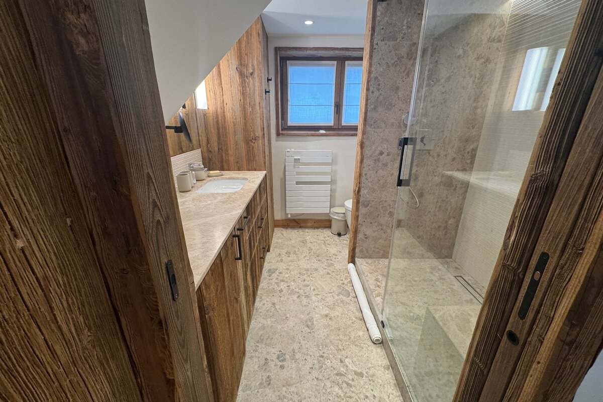 Rustic bathroom with warm wooden walls & cabinetry, a marble vanity, walk-in shower, and window, creating a cozy, natural feel.