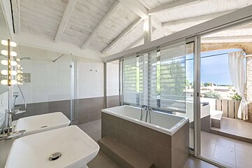 Bright, modern bathroom with a white tub, glass shower, dual sinks, vaulted white wooden ceiling, and sliding doors opening to a sunny outdoor terrace.