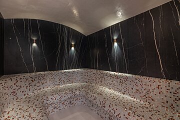 A luxurious steam room features black marble walls with white veins, curved mosaic-tiled benches, and modern wall sconces providing soft light.