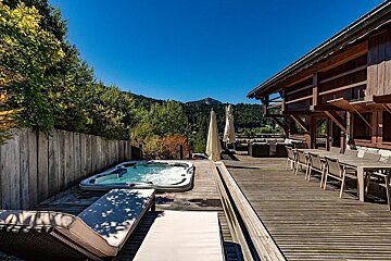 A spacious wooden deck features a hot tub, sun loungers, outdoor dining, and seating, set against a forested mountain view under a clear blue sky.