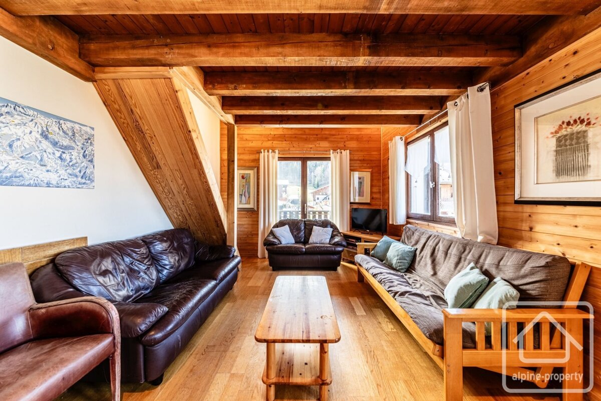 A warm, wood-paneled chalet living room with exposed beams, leather and futon sofas, a wooden coffee table, and bright windows.