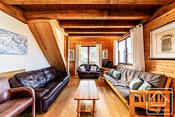 A warm, wood-paneled chalet living room with exposed beams, leather and futon sofas, a wooden coffee table, and bright windows.