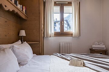 Cozy alpine bedroom with wood-paneled walls, a bed with an open book, and a window showing a mountain chalet view.