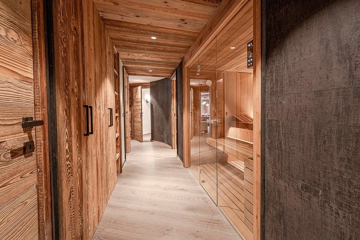 Modern wooden hallway with warm timber walls and ceiling, light floor, and a stylish glass-fronted sauna visible on the right side.