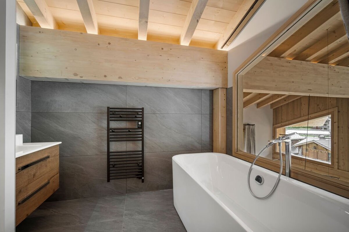 Modern chalet bathroom featuring a freestanding white tub, grey stone tiles, and light wood beams.
