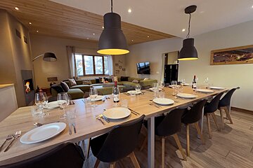 A cozy, modern dining room with a long table set for dinner, adjacent to a living area with a large green sofa, TV, and a lit fireplace.