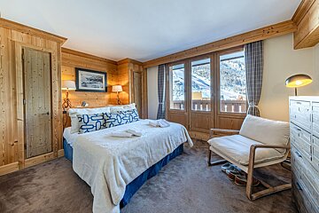 A cozy chalet bedroom with wood paneling, a large bed, and a balcony view of snowy mountains. Features an armchair and a unique antler-themed lamp.