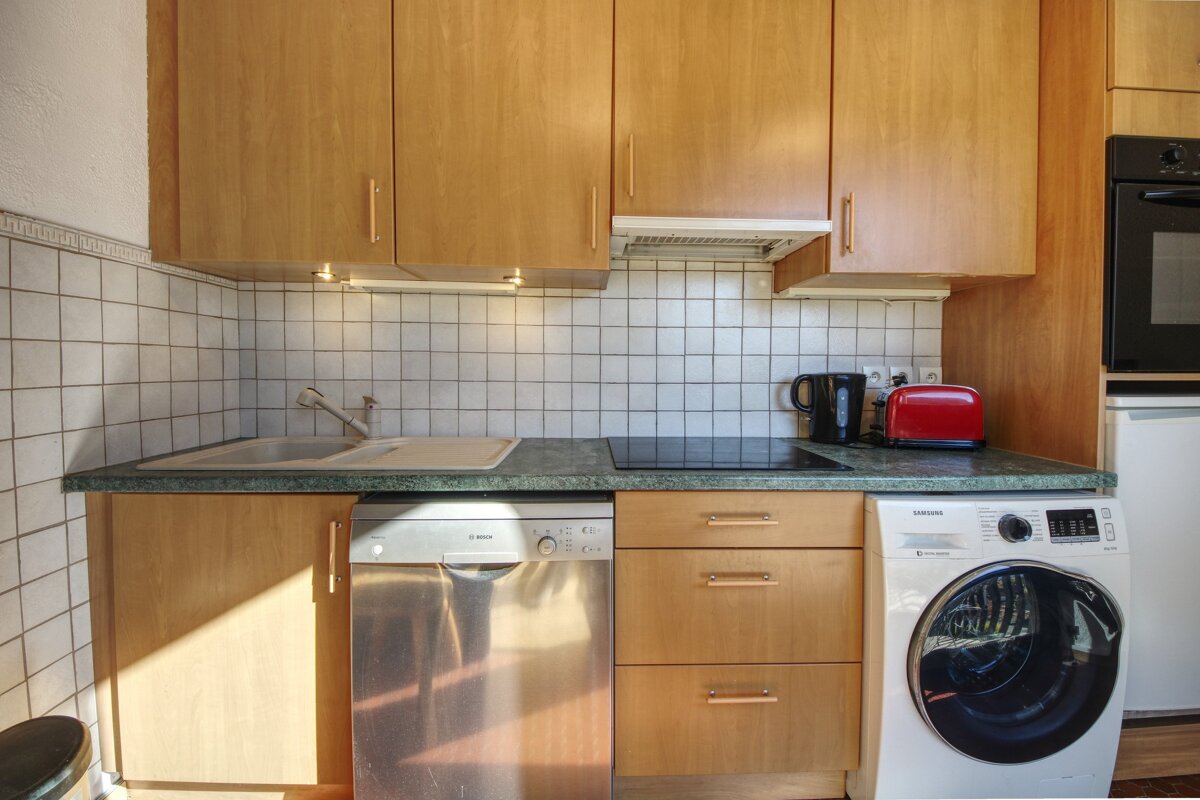 A modern kitchen with light wooden cabinets, a white tiled backsplash, and a green countertop. Appliances include a sink, stove, dishwasher, washing machine, and oven.