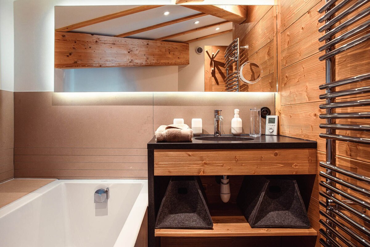 A warm, modern bathroom with extensive wood paneling, featuring a bathtub, a dark vanity with a sink, and an illuminated mirror. Rustic alpine style.