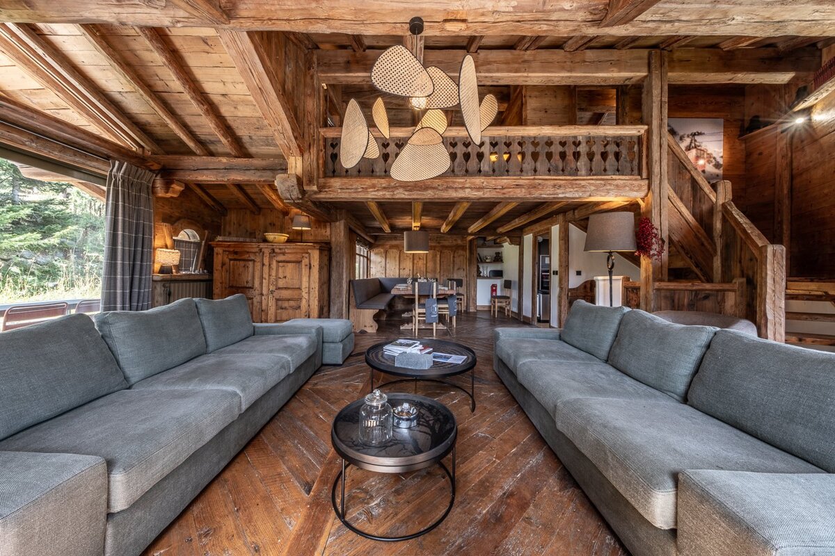 Cozy, rustic wooden chalet living room with exposed beams, two long grey sofas, round coffee tables, and a unique modern light fixture.