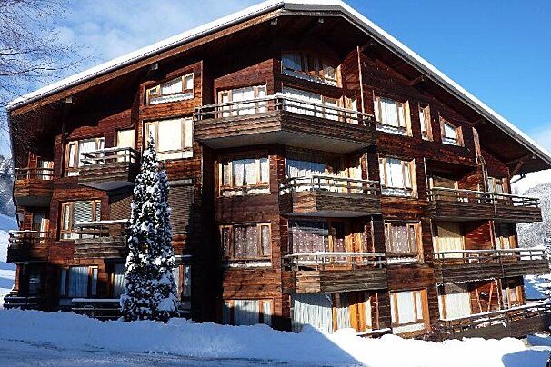 A large, snow-covered wooden chalet with multiple balconies stands against a clear blue sky. A tall, snow-laden evergreen tree is in front.