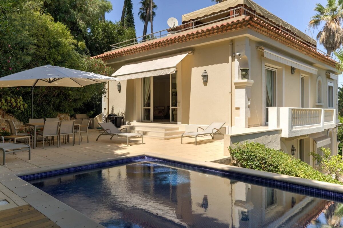 A cream-colored villa with a tiled roof overlooks a sparkling blue swimming pool and a sunny patio with outdoor dining and lounge chairs.